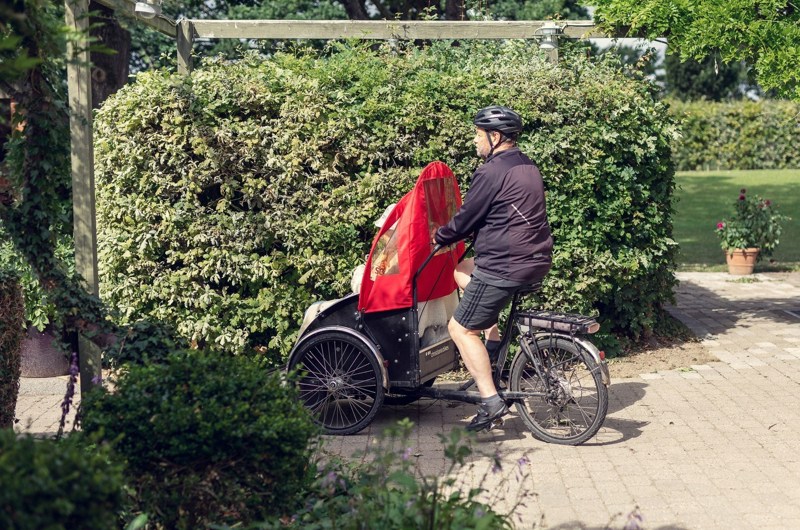 En frivillig på Rickshaw cykel med beboer på vej ud på en cykeltur. Grøn hæk i baggrunden.