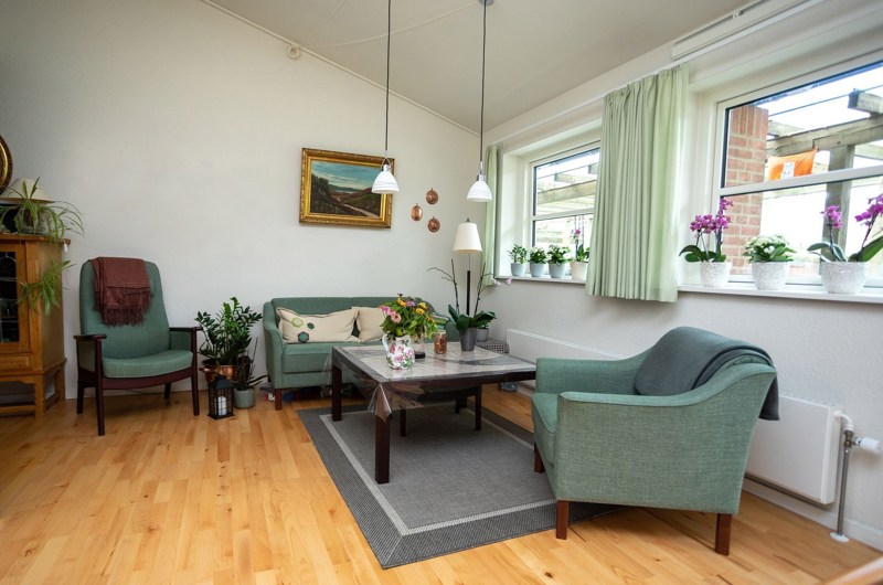 En bolig på Solgaven Næstved. En sofa, 2 stole i en grønlig farve samt et sofabord. Et tæppe under border, lamper over sofabord.2 vinduer med gardiner