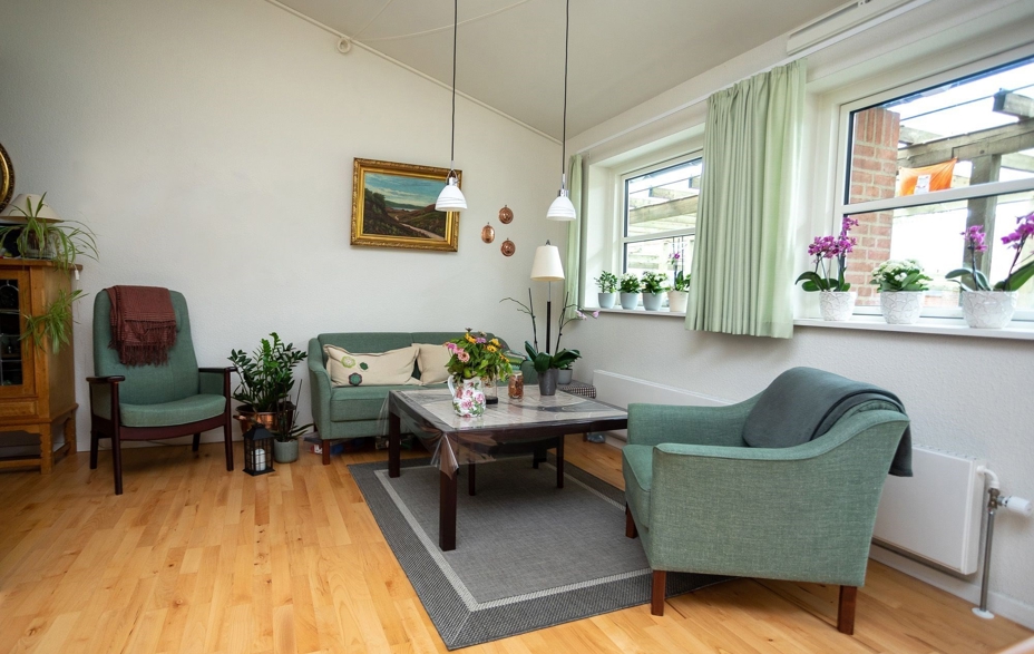 En bolig på Solgaven Næstved. En sofa, 2 stole i en grønlig farve samt et sofabord. Et tæppe under border, lamper over sofabord.2 vinduer med gardiner