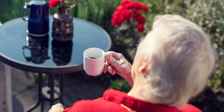 Beboer på Solgaven Næstved sidder udenfor og drikker kaffe ved rundt havebord med røde blomster i baggrunden. Der er også blomster i en vase på bordet.