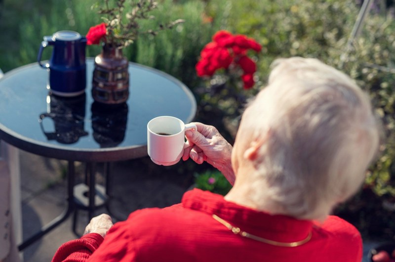 Beboer på Solgaven Næstved sidder udenfor og drikker kaffe ved rundt havebord med røde blomster i baggrunden. Der er også blomster i en vase på bordet.