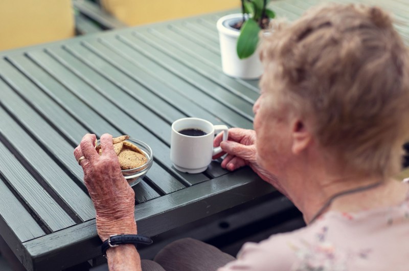 En beboer der sidder og nyder en kop kaffe og en småkage ved et grønt bord med en blomst på bordet i vinterhaven.
